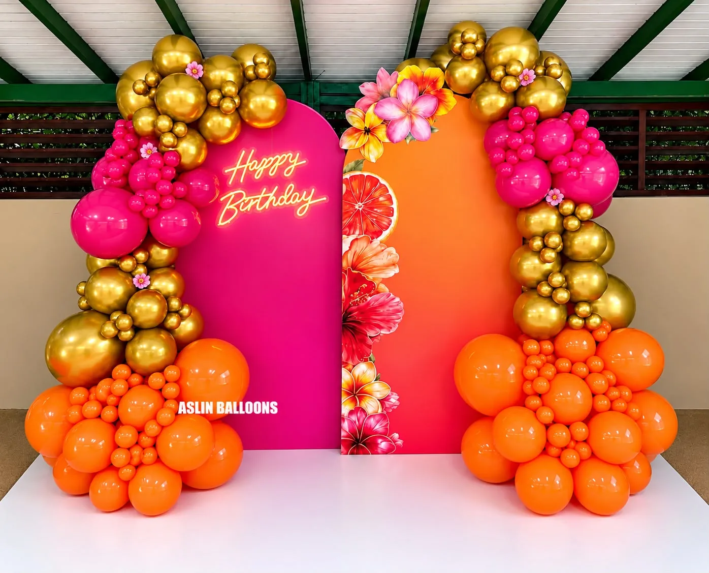 Decoración de cumpleaños en fucsia, naranja y dorado: paredes en arco impresas con flores tropicales, letrero de neón 'Happy Birthday' y arcos de globos.