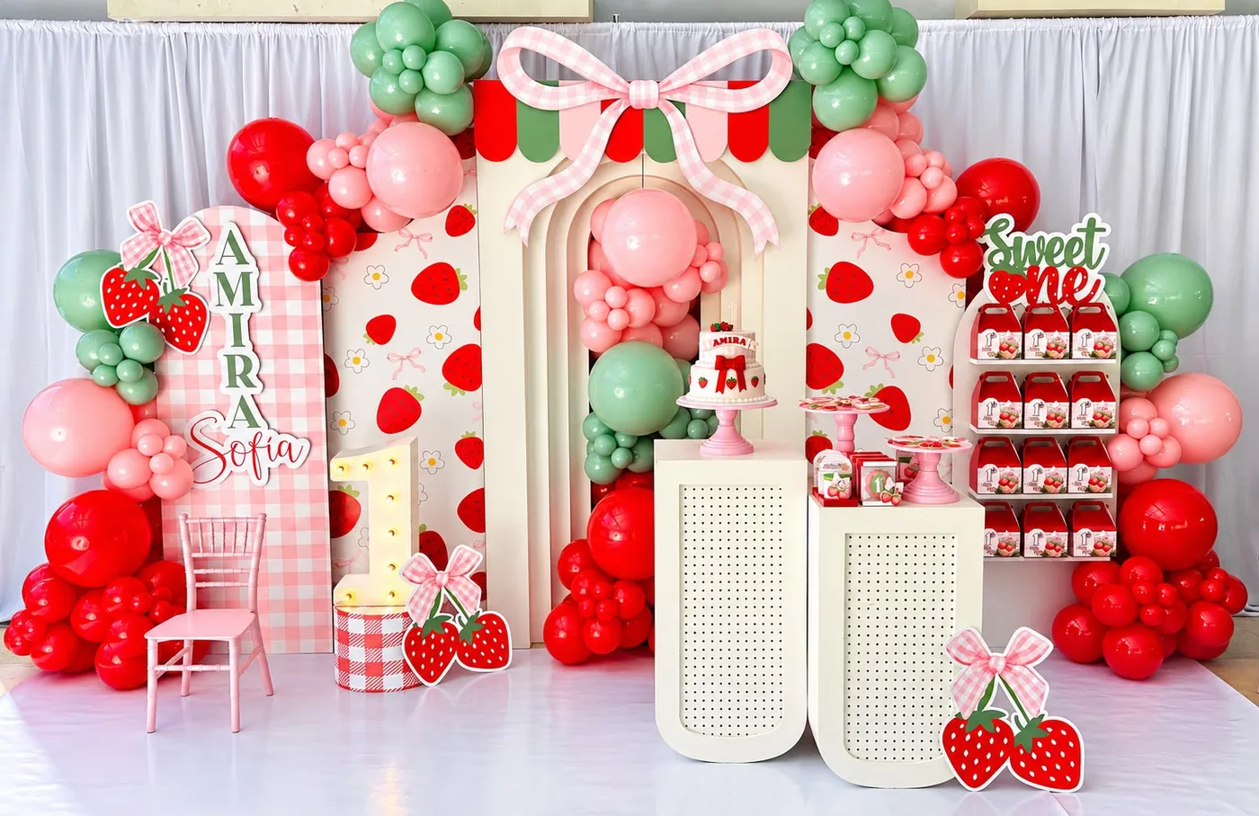 Decoración de primer cumpleaños con tema de fresas: panel en arco a cuadros rosa con nombre troquelado, paneles con fresas, lazo gigante, letrero 'Sweet One', figuras de fresas y globos rojos, rosa y verde.
