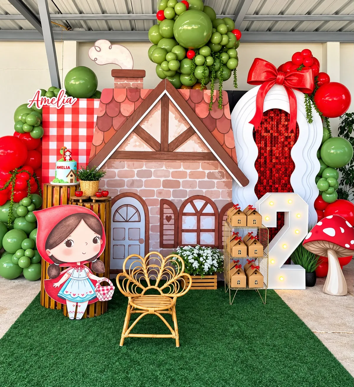Decoración de cumpleaños con tema de Caperucita: casita de campo impresa a tamaño real, figura troquelada de la niña, número 2 iluminado y globos verdes y rojos.