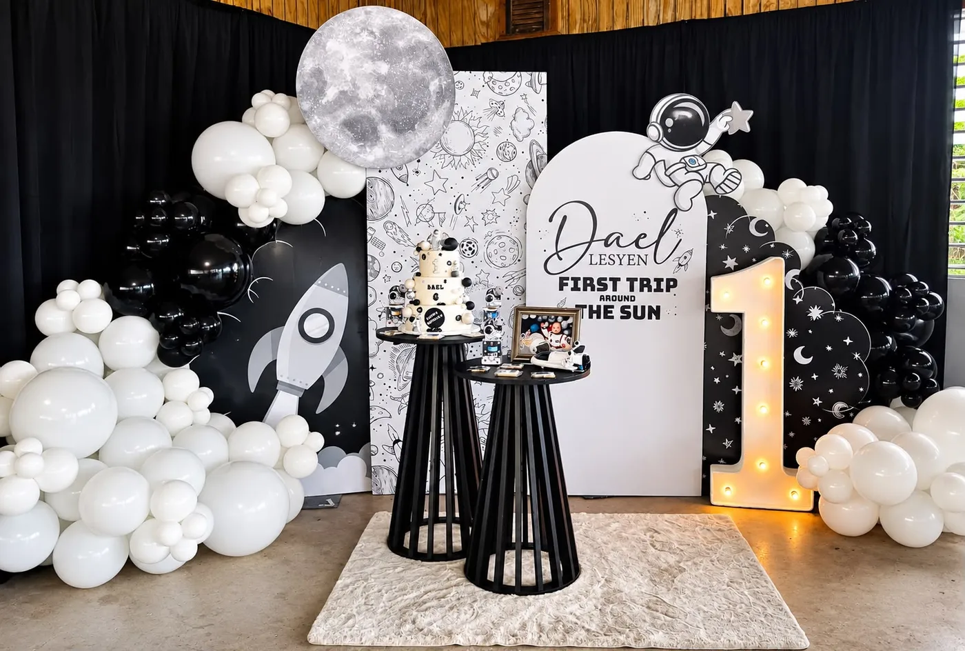 Decoración de primer cumpleaños con tema espacial: panel en arco con nombre y la frase 'First trip around the sun', figura troquelada de astronauta, luna, cohete, número 1 iluminado y globos blancos y negros.