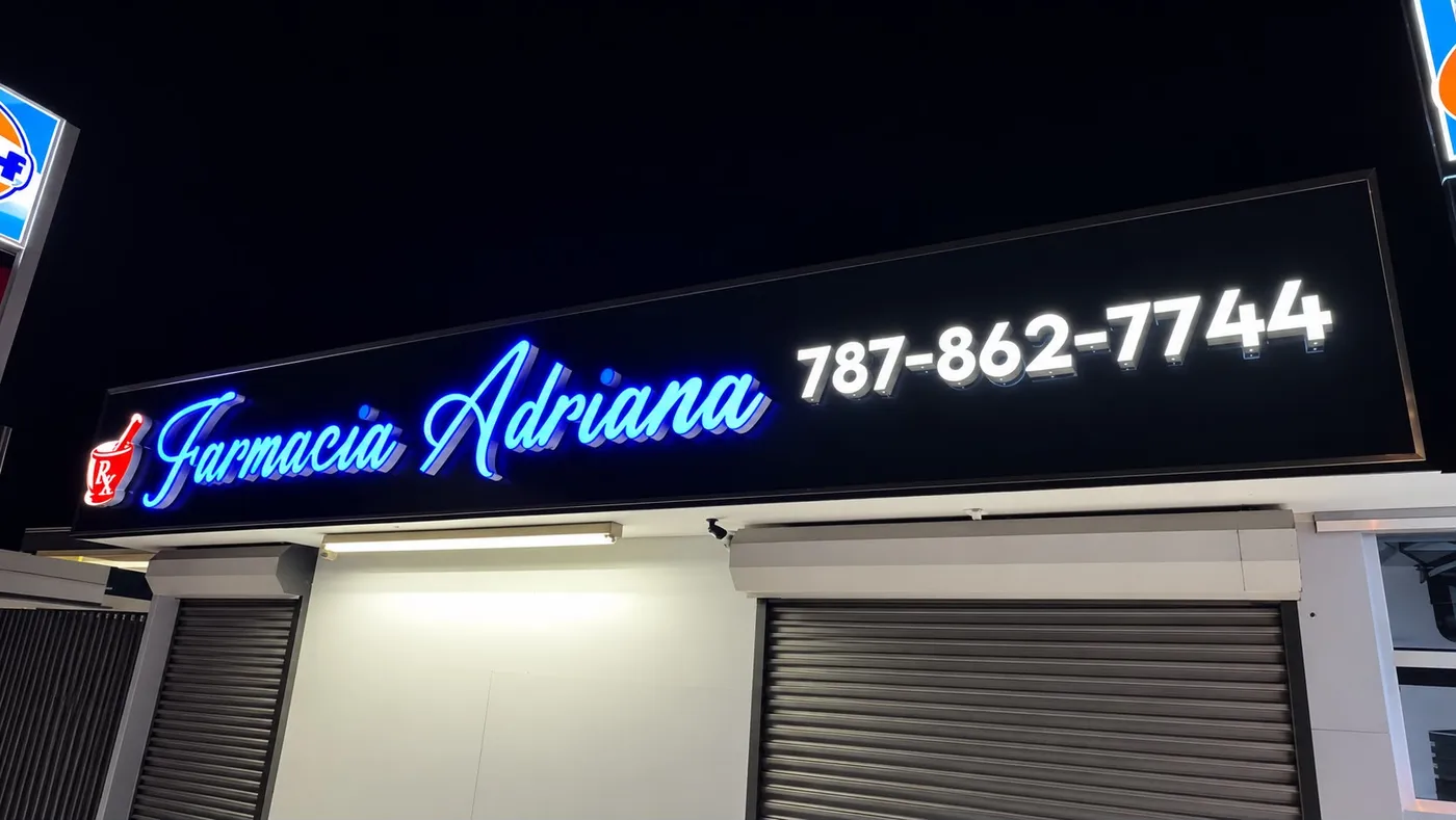 Cajuela lumínica de una farmacia encendida de noche, con el nombre en letras azules iluminadas y el teléfono en letras blancas.